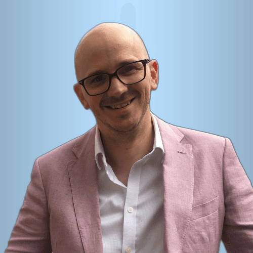 Headshot of Tim Jackson wearing a pink jackets, open necked white shirt and glasses. He is smiling and looking directly at the camera.